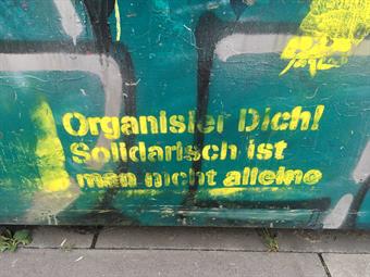 You can see a wall sprayed with Grafity. The background of the sprayed picture is turquoise with two letters on it in black/silver. Above the background image is a stencil "Organize yourself! In solidarity you're not alone" in bright yellow writing sprayed over the background picture. 